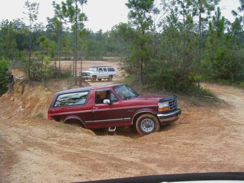 1990 Ford Bronco - A red 1992 Ford Bronco is partially submerged in mud on an off-road trail, with another SUV in the background.