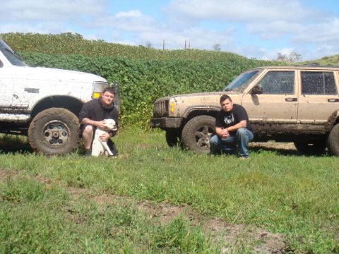 1992 Ford Bronco - Two men and a dog stand next to two muddy off-road vehicles in a grassy field.