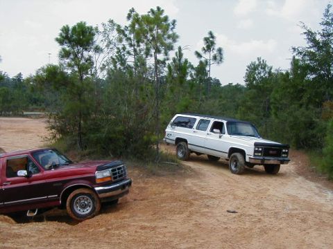 1990 Ford Bronco - Two trucks navigate a sandy off-road trail surrounded by trees.
