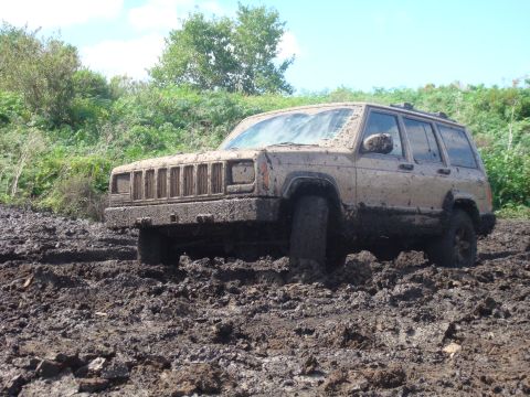 1992 Ford Bronco - A mud-splattered Jeep Cherokee XJ struggles in deep mud, its tires submerged.