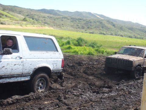 1992 Ford Bronco - Two off-road vehicles are stuck in thick mud on a scenic, grassy hillside.