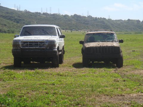 1992 Ford Bronco - Two off-road vehicles, a white 1992 Ford Bronco and a brown Jeep Cherokee, sit covered in mud after an outing.