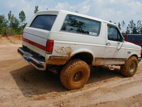 1990 Ford Bronco - A muddy white Ford Bronco navigates a dirt track, with another vehicle partially visible in the background.