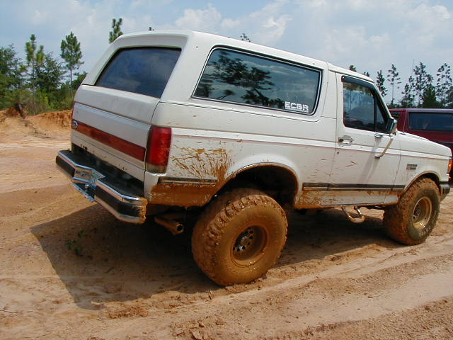 White Ford Bronco covered in mud, seen from the rear-right at a 3/4 angle on a dirt track