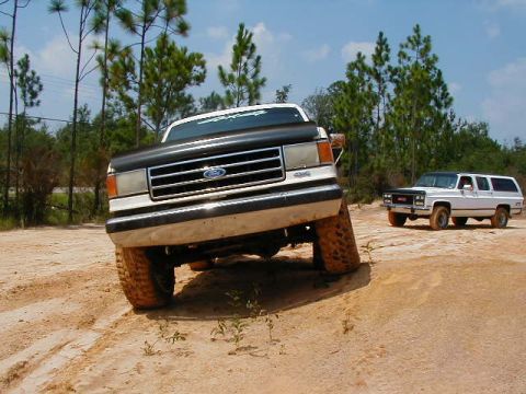 1990 Ford Bronco - Two SUVs, a Ford Bronco and a GMC Suburban, navigate a sandy off-road trail.
