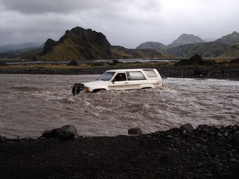 1996 Ford Bronco - A white SUV navigates a fast-flowing river in a , mountainous landscape under an overcast sky.