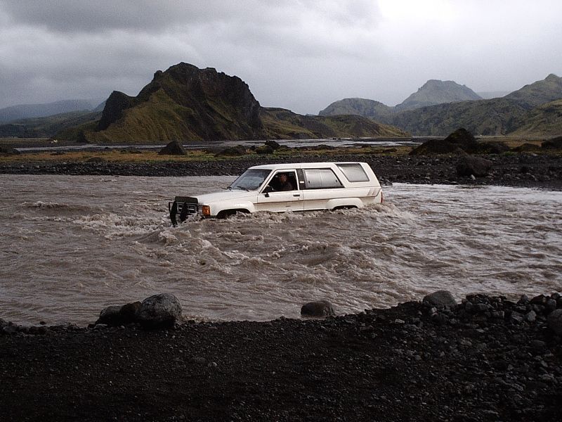 White SUV fording a muddy river with mountains behind under overcast sky