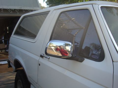 1992 Ford Bronco - A white SUV is parked in front of a garage. The side mirror reflects a person wearing a red shirt.