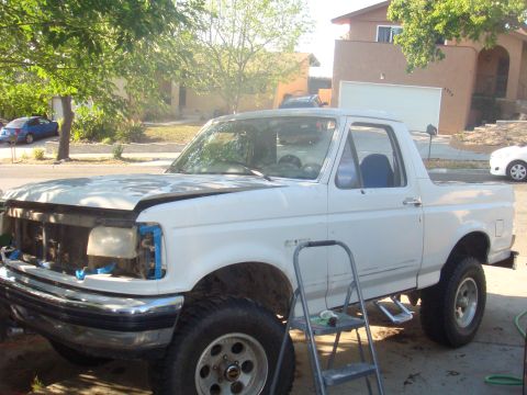 1992 Ford Bronco - A white 1992 Ford Bronco is parked outside a house, undergoing some work with its hood open and headlights removed.