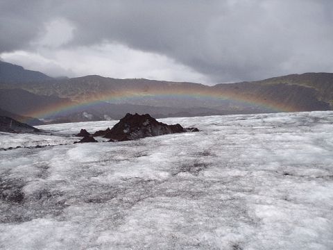 1996 Ford Bronco - A rainbow arcs over a vast glacier, with dark rocky outcrops breaking the icy expanse under a dramatic sky.