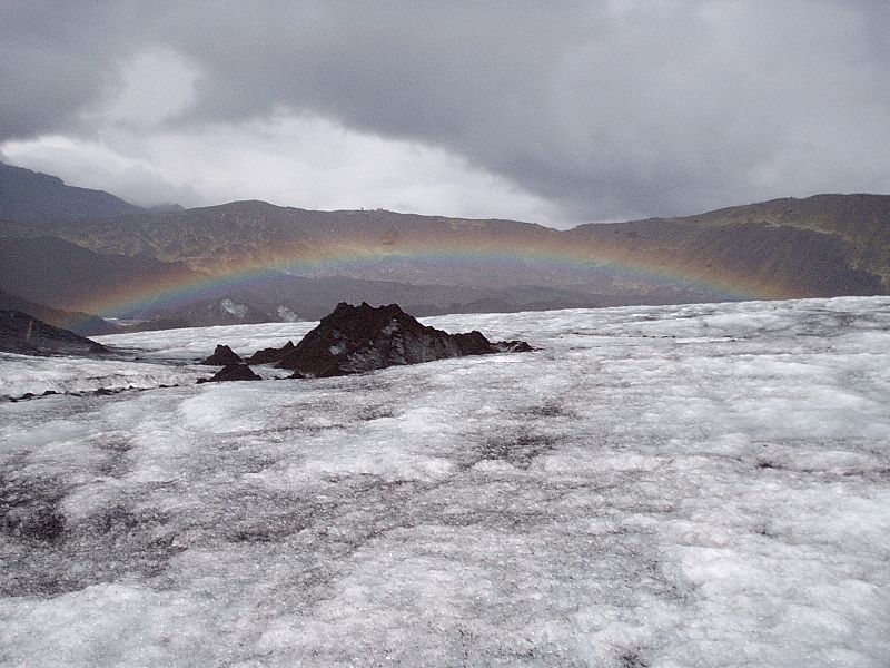Rainbow over rocky outcrop on a glacier under cloudy mountains