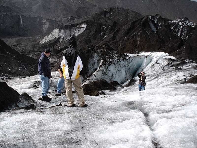 People standing on an ash-covered glacier with blue ice and mountains