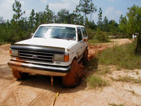 1990 Ford Bronco - A white 1990 Ford Bronco is stuck in the mud on a dirt road, with trees and another vehicle in the background.