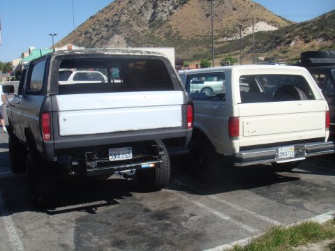 1992 Ford Bronco - Two Ford Broncos, one cream-colored and one gray, are parked in a lot, both showing their rear ends.