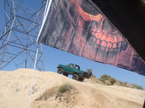 1992 Ford Bronco - A green off-road truck navigates a sandy incline, partially obscured by a large flag with a skull graphic.