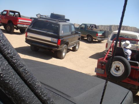 1992 Ford Bronco - A group of off-road vehicles, including a black 1992 Ford Bronco, navigate a sandy terrain under a clear blue sky.