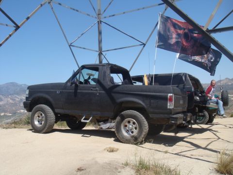 1992 Ford Bronco - A black 1992 Ford Bronco is parked near another vehicle under a large metal structure, with skull-themed flags waving in the breeze.