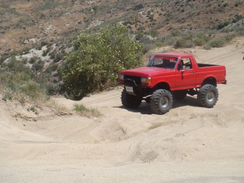 1992 Ford Bronco - A red lifted truck with large off-road tires navigates a sandy, hilly terrain.