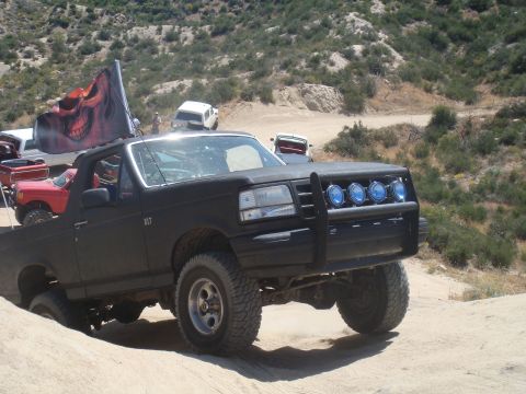 1992 Ford Bronco - This black 1992 Ford Bronco is equipped with off-road tires and a skull flag, navigating a dirt trail.