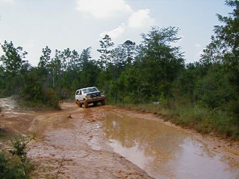 1990 Ford Bronco - A white 1990 Ford Bronco tackles a challenging muddy off-road course, splashing through a large water obstacle.