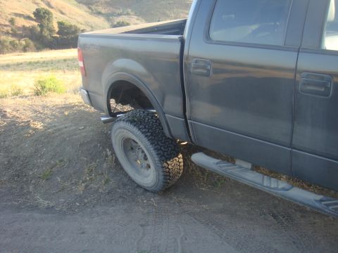1992 Ford Bronco - A dark gray pickup truck is shown from the rear quarter angle, parked on a dirt road with hills in the background.