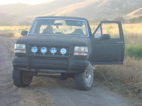 1992 Ford Bronco - This black Ford Bronco is parked on a dirt road with its driver's door open, facing the camera with hills in the background.
