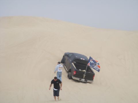 1992 Ford Bronco - A black pickup truck is stuck in the sand dunes, with two people approaching it. A flag is mounted on the back of the truck.
