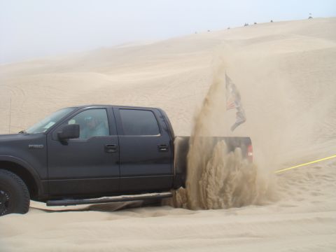 1992 Ford Bronco - A black pickup truck is driving through sand dunes, creating a large plume of sand.