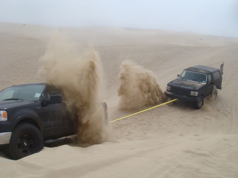 1992 Ford Bronco - A black 1992 Ford Bronco is being towed by a black Ford F-150 through sand dunes, kicking up dust.