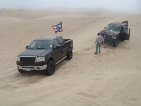 1992 Ford Bronco - Two trucks are in the sand dunes, one is stuck and being pulled by the other.