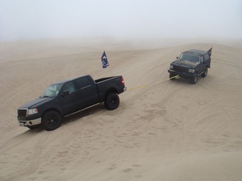 1992 Ford Bronco - A black Ford F-150 tows a black Ford Bronco through sandy terrain under a foggy sky.