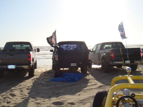 1992 Ford Bronco - Three pickup trucks are parked on a beach, with two flying flags and a dune buggy visible in the foreground.