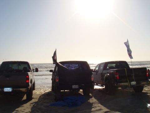 1992 Ford Bronco - Multiple vehicles parked on the sand near the ocean waves under a bright sky.