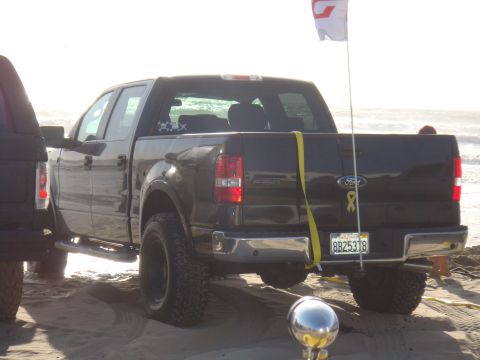 1992 Ford Bronco - A dark Ford F-150 pickup truck is parked on the sand near the ocean, with a flag on a pole in the bed.