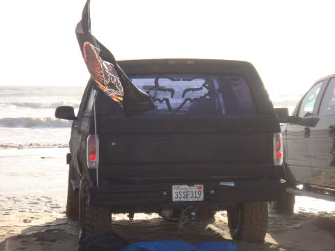 1992 Ford Bronco - A black 1992 Ford Bronco is parked on a sandy beach, displaying a flag and a Fox Racing decal on its rear window.