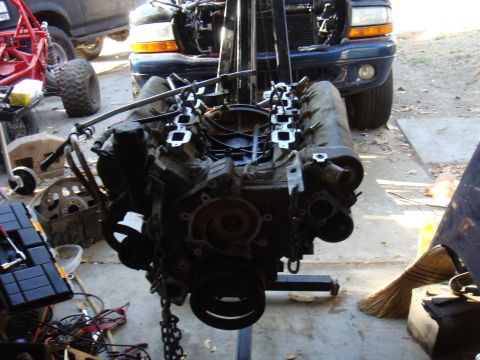 1992 Ford Bronco - An engine is suspended by a hoist, with a dark blue truck partially visible behind it.
