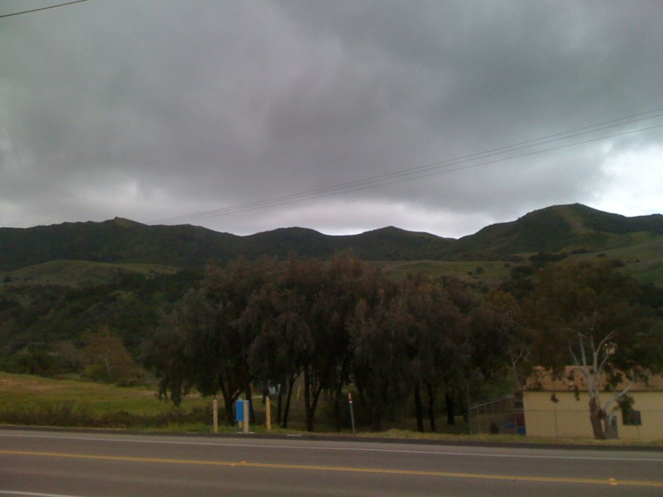 road, trees, and hills under cloudy sky. Building on right
