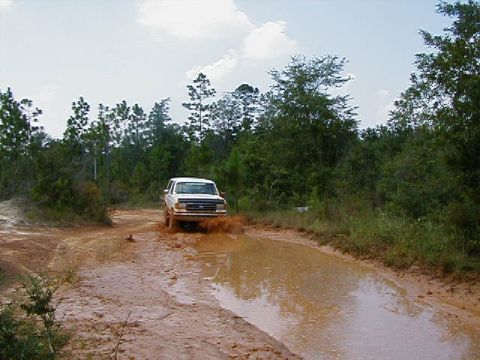 1990 Ford Bronco - A white pickup truck splashes through a muddy track in a wooded area.