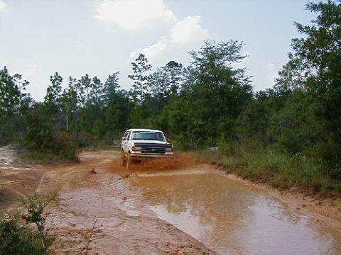 1990 Ford Bronco - A white 1990 Ford Bronco navigates a muddy path, kicking up water and dirt.