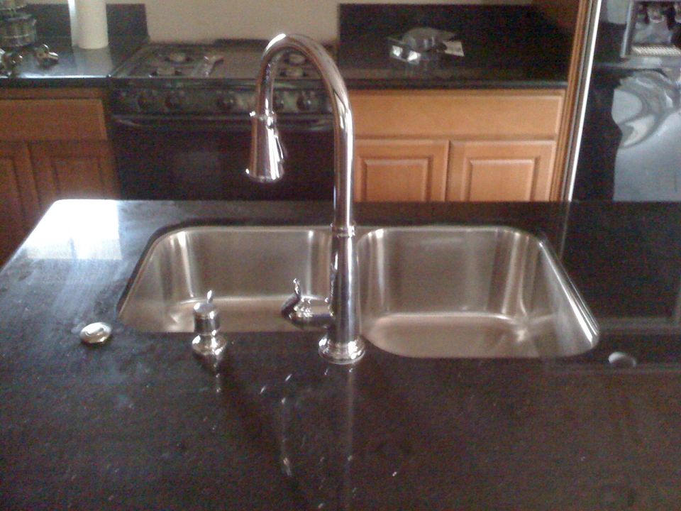 Double basin stainless steel kitchen sink with chrome faucet and soap dispenser on granite countertop. Stove and cabinets in background