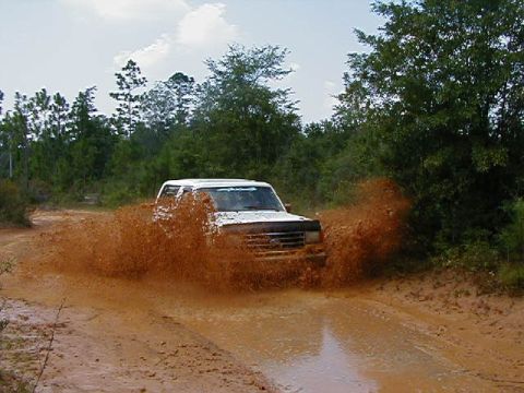 1990 Ford Bronco - A white SUV navigates a muddy trail, kicking up a large spray of dirt and water.