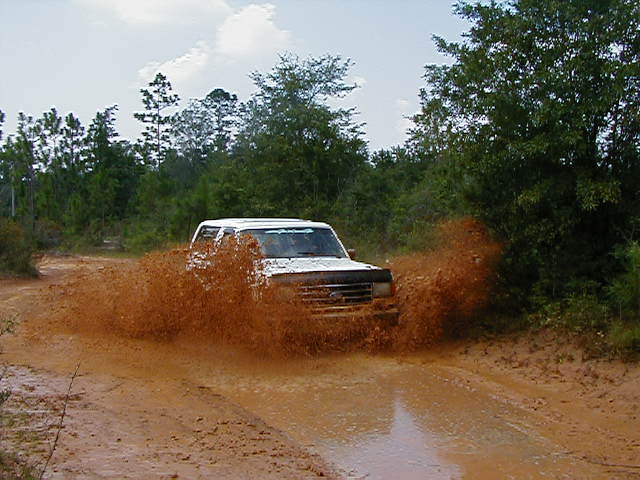 White SUV splashing through a muddy puddle on a dirt road, trees and sky in background