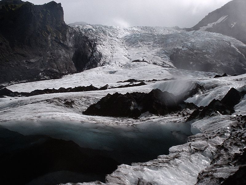 Glacier with meltwater pools and rocky ridges under misty mountains