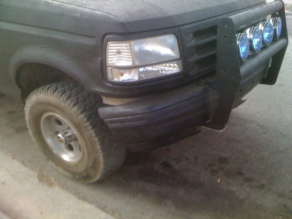 1992 Ford Bronco front end, showing headlight, grille, and aftermarket bumper with four lights. Tire and fender visible