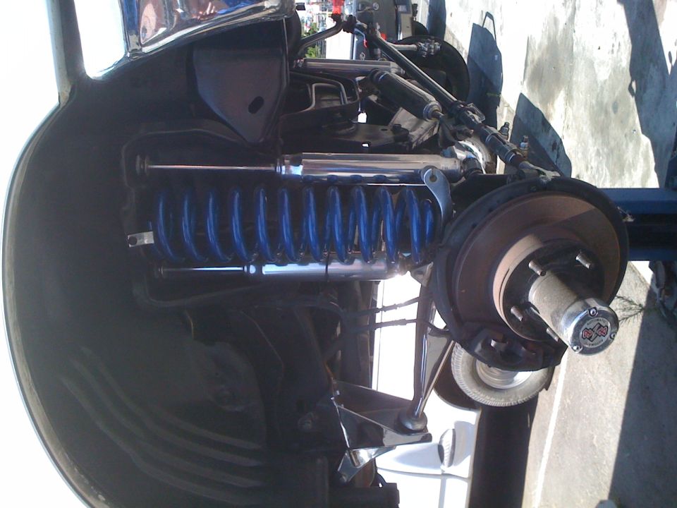 Close-up of a vehicle's front suspension with blue coil springs and a chrome shock absorber. The wheel hub and brake rotor are visible