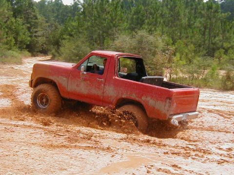 1990 Ford Bronco - This red SUV is struggling to get through a muddy off-road trail, with its tires submerged in thick mud.