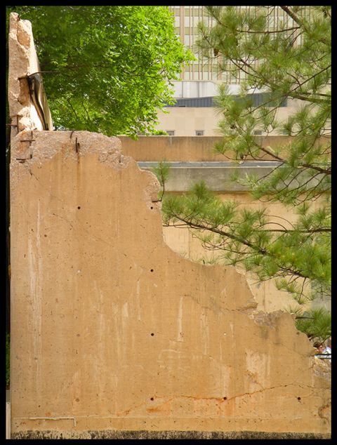 1998 Subaru Legacy - A section of a damaged concrete wall with visible rebar, set against a backdrop of trees and urban buildings.