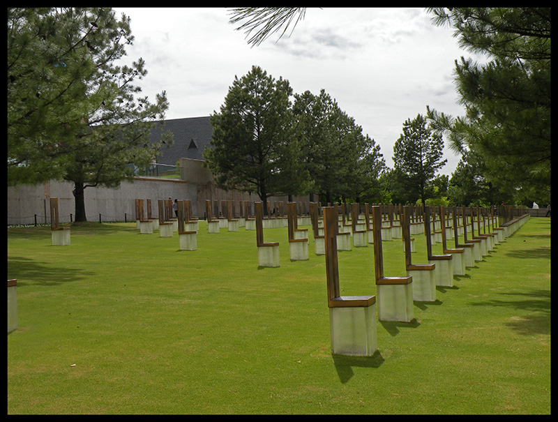 Rows of empty chairs on concrete bases in a grassy field, with trees and a building in the background