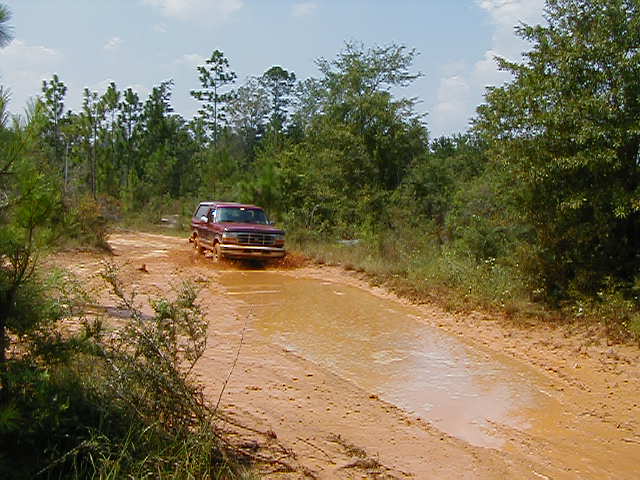 Red 1990 Ford Bronco driving through a muddy puddle on a dirt road, trees and sky in background