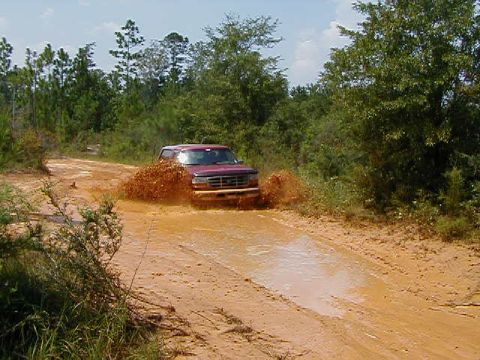 1990 Ford Bronco - This SUV is navigating a challenging muddy path, kicking up a large spray of mud.